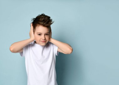 Sinsi yaramaz çocuk gülümsüyor ve elleriyle kulaklarını kapatıyor. Duymak ve itaat etmek istemediğini gösteriyor. Açık mavi üzerinde izole edilmiş bir stüdyo portresi