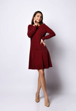 Uzun kollu, zengin kırmızı bir elbisesi ve çıplak ayakkabısı olan çapraz bacaklı eli kalçalı, beyaz tenli, çapraz bacaklı genç bir kadın.