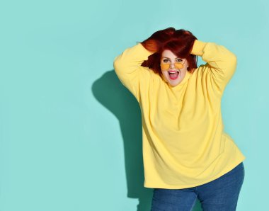 Uzun kızıl saçlarının arasında neşeli bir şekilde parmaklarını gezdiren kilolu, tatlı genç bir kadın. Mavi üzerine üç çeyrek boy atış.