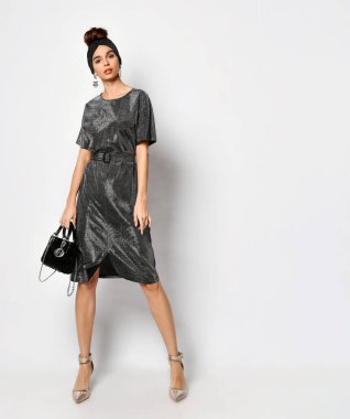 Zarif, parlak, kömür rengi bir elbise giymiş, elinde siyah bir çantayla duran alımlı genç bayan. Tam uzunluktaki stüdyo çekimi beyazda izole edildi