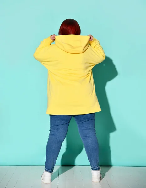 Back view of a voluptuous lady in jeans and bright yellow hoodie putting capuche on her head. — Stock Photo, Image