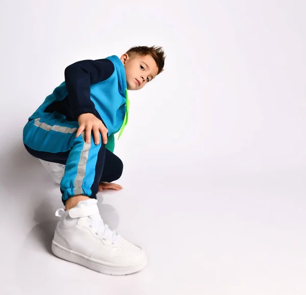Kis fiú színes melegítőben, tornacipőben. Gyakorlatokat végez, vagy a lábánál rúg, fehér alapon elszigetelve pózol. — Stock Fotó