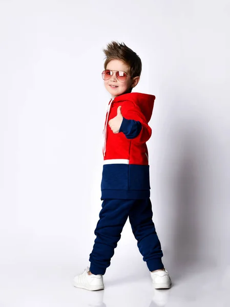 Kleiner blonder Junge in blauem und rotem Trainingsanzug, Turnschuhen, Sonnenbrille. Lächeln dreht sich um, zeigt den Daumen nach oben, posiert isoliert auf weiß — Stockfoto