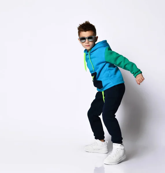 Little kid in sunglasses, colorful sport suit and sneakers. He performing a kick by his leg, posing isolated on white — Stock Photo, Image