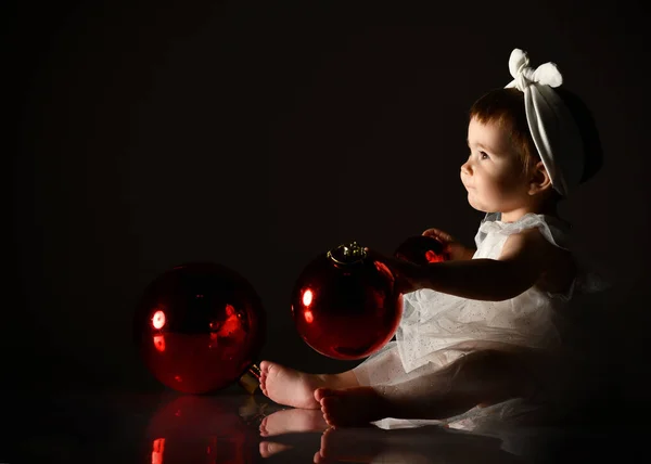 Pequena fêmea de cabeça branca e vestido, descalça. Ela segurava duas bolas vermelhas, sentada no chão. Crepúsculo, fundo preto. — Fotografia de Stock