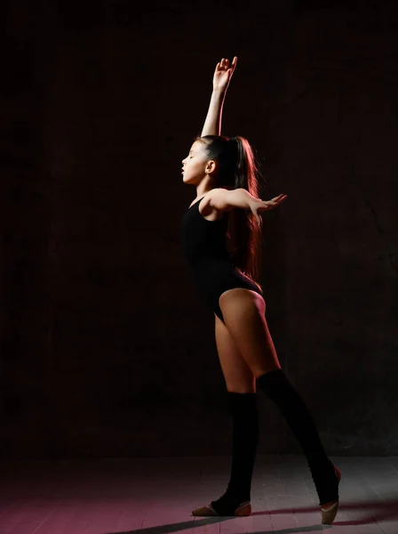 Giovane ragazza ginnasta in nero sport corpo e speciale tomaie in piedi e fare ginnastica posa su sfondo scuro — Foto Stock