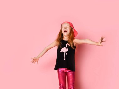 Şapkalı, siyah bluzlu ve taytlı heyecanlı sarışın. Kollarını araladı ve sevinçle çığlıklar attı. Pembe bir stüdyo arka planına karşı poz verdi..