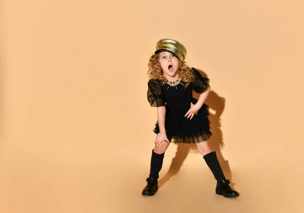 Hübsches kleines Mädchen in schwarzem Kleid, goldener Mütze auf lockigem Haar und schwarzen Stiefeln singt ein Lied, das vorwärts gebeugt mit der Hand auf dem Knie, isoliert auf Pfirsichhintergrund — Stockfoto