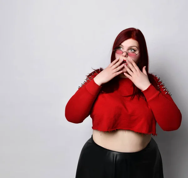 Plus size ginger girl in red top, black skirt, sunglasses. Covered mouth with hands, looking scared, posing isolated on white
