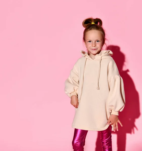 Симпатична маленька дівчинка з підтягнутим позуванням з впевненим виглядом в блідо-рожевому светрі і глянцевих легінсах. Три чверті довжини ізольовані на рожевому, копіювати простір — стокове фото