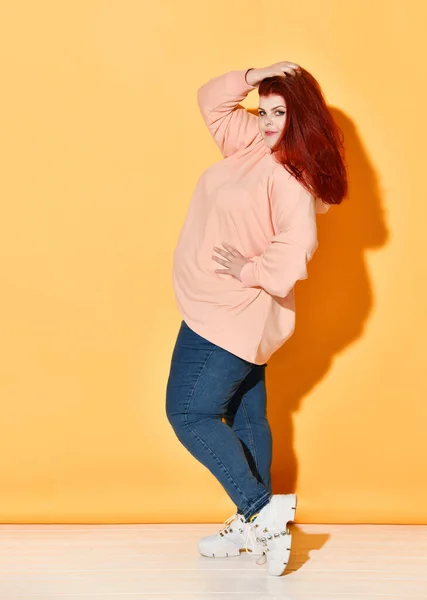 Joyful red-haired curvy model winking and pointing aside showing tongue tip, joking about someone. Full length shot isolated on orange.