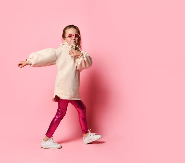 Чарівна маленька дівчинка в окулярах, теплий блідо-рожевий светр і легінси йде з легкою посмішкою. Повнометражний знімок ізольовано на рожевому, простір копіювання — стокове фото