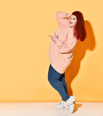 Neşeli kızıl saçlı kıvrımlı model göz kırpıyor ve dil ucu gösteriyor, biri hakkında şaka yapıyor. Turuncuda tam boy atış izole.