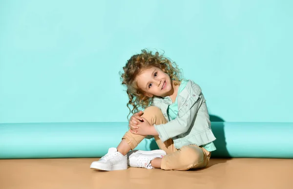 Linda niña rubia con ropa casual se sienta en el suelo abrazando la rodilla, inclinando la cabeza y sonriendo. Retrato de estudio sobre fondo turquesa — Foto de Stock