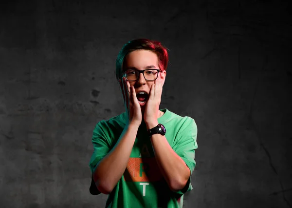 Joven adolescente positivo en camiseta verde y gafas de pie y expresando malentendido sobre fondo oscuro — Foto de Stock