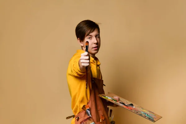Νεαρός έφηβος ζωγράφος αγόρι σε κίτρινο hoodie και jumpsuit με καβαλέτο δείχνει βούρτσα του στην κάμερα πάνω από κίτρινο φόντο — Φωτογραφία Αρχείου