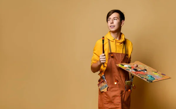 Νεαρός έφηβος ζωγράφος αγόρι σε κίτρινο hoodie και jumpsuit στέκεται και κρατώντας καβαλέτο, χρώμα και πινέλο στα χέρια πάνω από το κίτρινο φόντο — Φωτογραφία Αρχείου
