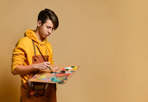 Joven adolescente pintor con capucha y mono amarillo de pie y pintando sobre caballete sobre fondo amarillo — Foto de Stock