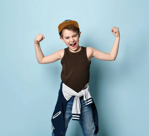 Adolescente masculino en gorra y ropa casual con estilo. Apretó los puños mostrando músculos y gritando, posando sobre fondo azul —  Fotos de Stock