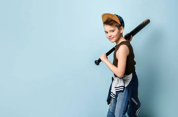 Sportief jongetje in mouwloos shirt, spijkerbroek en pet klaar om te slaan met honkbalknuppel vanaf zijn schouder. Zijaanzicht schot geïsoleerd op blauwe achtergrond — Stockfoto