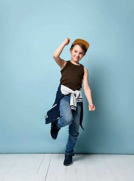 Teenager v čepici, hnědé tričko, džíny, kapuci přivázanou kolem pasu a tenisky. Usmívá se, pózuje na modrém pozadí — Stock fotografie