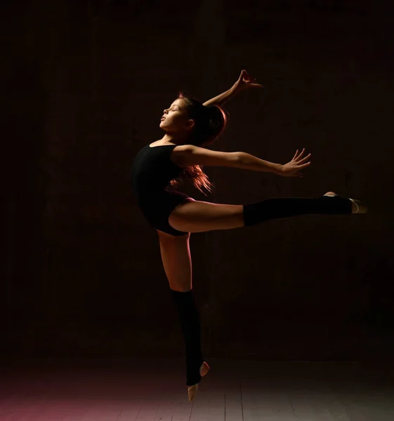 Young girl gymnast in black sport body and special uppers jumping in gymnastic pose over dark background — Stock Photo, Image