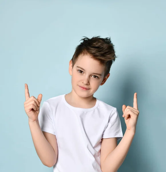 Bel bambino amabile in t-shirt bianca che guarda avanti con il sorriso mettendo entrambe le dita indice in su. Ritratto a mezza lunghezza isolato su azzurro Foto Stock