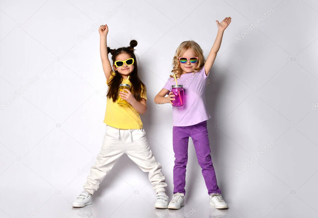 Little females in sunglasses, colorful casual clothes. Holding cocktail bottles, smiling, raised hands, posing isolated on white