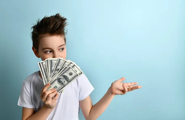 Tarz sahibi çocuk nakit dolarları elinde tutuyor nasıl yatırım yapacağını ya da nasıl daha iyi harcayacağını merak ediyor. Açık mavi üzerine izole edilmiş yarım boy portre Stok Resim