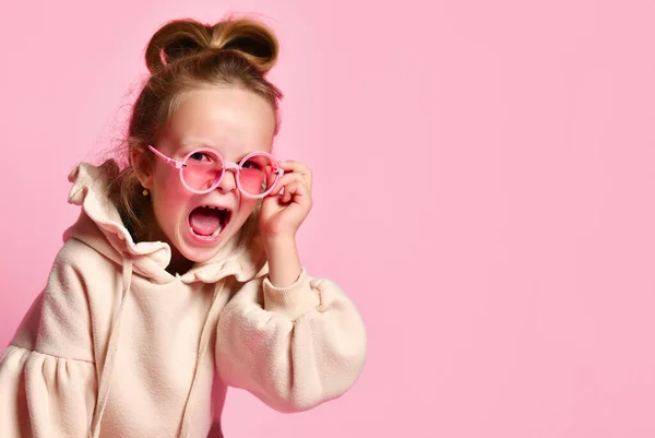 Неслухняна маленька дівчинка з високим волоссям, одягнена в рожевий светр, кричить сердито розкладаючи окуляри на обличчі. Крупним планом знімок ізольовано на рожевому, місце для копіювання — стокове фото