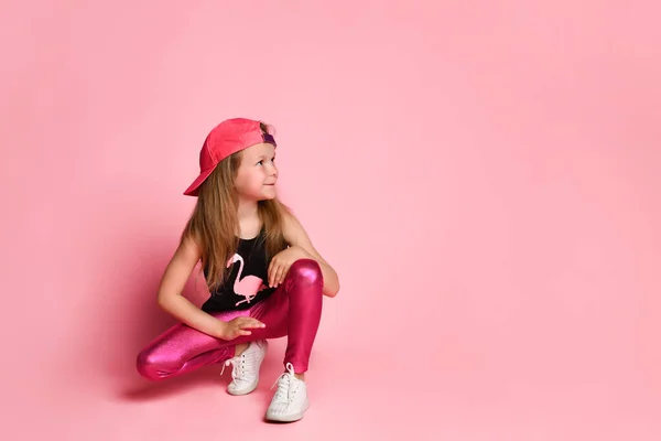 Charmant actief kleuter meisje in een trendy modieuze outfit, speels en lachend, hurkend en kijkend naar de zijkant op een roze achtergrond — Stockfoto