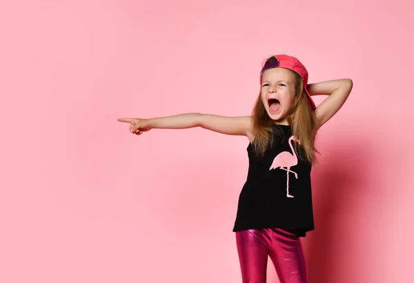 Kind in pet, blouse met bedrukte flamingo en legging. Ze schreeuwde en wees naar de zijkant door wijsvinger, poserend op een roze achtergrond. Sluiten. — Stockfoto