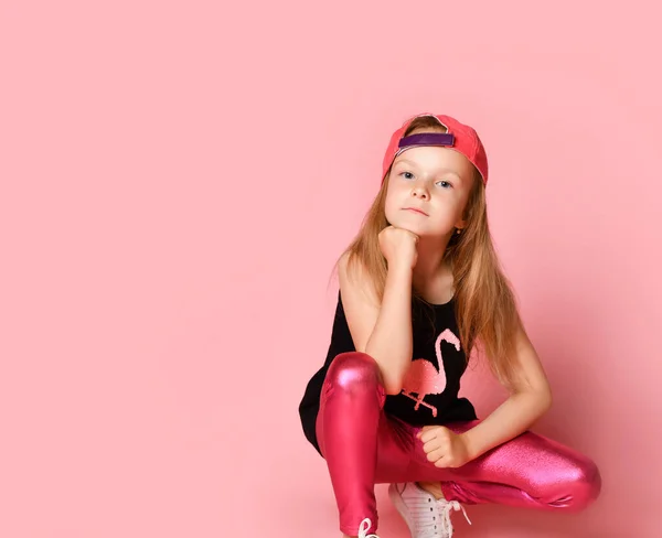 Charmant actief kleuter meisje in een modieuze modieuze jurk, speels en lachend, hurkend, naar je kijkend met haar vuist op haar hoofd. op een roze achtergrond. — Stockfoto