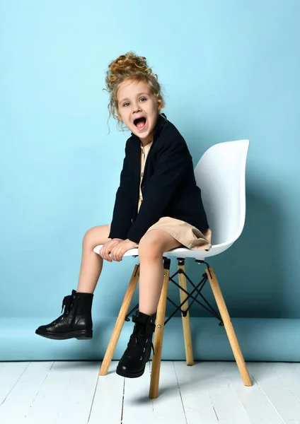 Niña en vestido beige, chaqueta negra y botas. Ella está gritando, sentada en una silla blanca sobre fondo azul del estudio — Foto de Stock