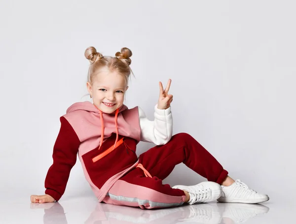 Affascinante bambina in bella tuta sportiva alla moda seduta sul pavimento mostrando due dita nel segno della vittoria. Ritratto ravvicinato isolato su grigio chiaro — Foto Stock