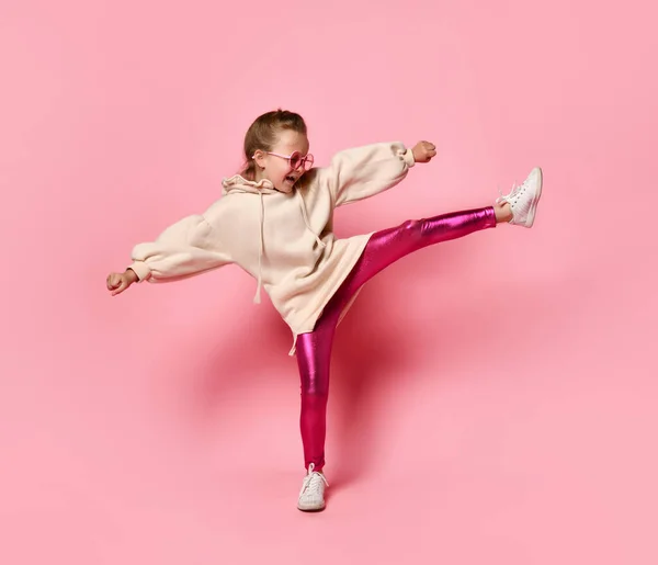 Zijaanzicht van een klein meisje met een capuchon, sprankelende benen en glazen die op een been staan en een ander been naar voren strekken. Studio schot geïsoleerd op roos — Stockfoto