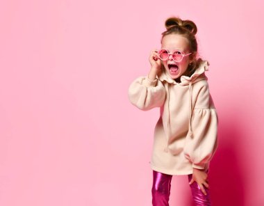 Komik saç stili olan, pembe kazak giyen, bağıran, suratında kızgın gözlük takan yaramaz bir kız. Yakın çekim pembe üzerine izole edilmiş, duygular.