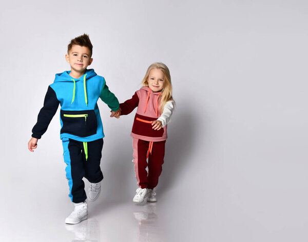 Мальчик и девочка в ярких спортивных костюмах идут вперед рука об руку. Полноразмерный портрет, изолированный на светло-сером, копировальное пространство
