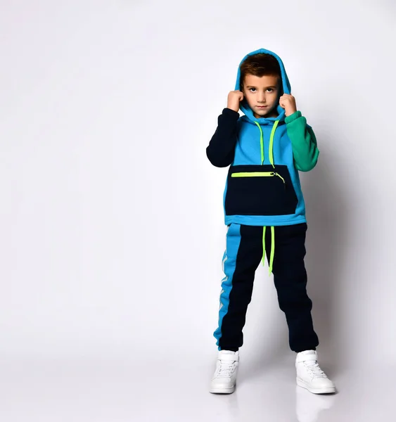 Niño pequeño en traje deportivo colorido y zapatillas de deporte. Se ha puesto la capucha mientras posaba aislado sobre fondo blanco del estudio. —  Fotos de Stock
