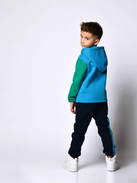 Αγόρι με πολύχρωμο αθλητικό κοστούμι, αθλητικά παπούτσια. Κοιτάζει πίσω, ποζάρει πίσω στην κάμερα, απομονωμένος σε λευκό στούντιο. — Φωτογραφία Αρχείου