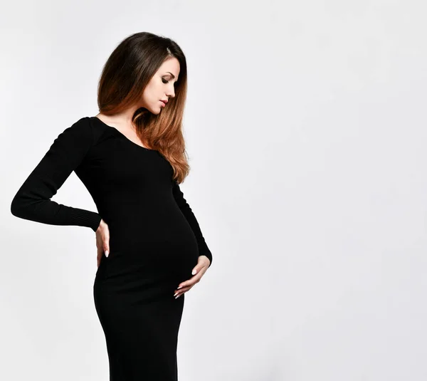 Siyah dar elbiseli esmer hamile kadın. O, beyaz üzerinde yanlamasına poz verirken elini karnının üzerine koydu. Kapat.