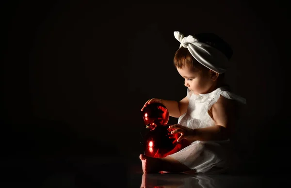 Niño pequeño con diadema blanca y vestido, descalzo. Ella jugando con dos bolas rojas, sentada en el suelo. Crepúsculo, fondo negro. — Foto de Stock