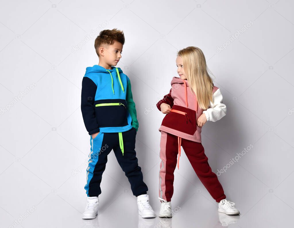 Little kids, boy and girl, in colorful tracksuits and sneakers. They posing isolated on white studio background. Hands in pockets