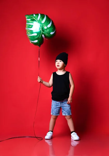 Piccolo ragazzo positivo in elegante abbigliamento casual e cappuccio in piedi e tenendo foglia stampato palloncino in mano su sfondo muro rosso — Foto Stock