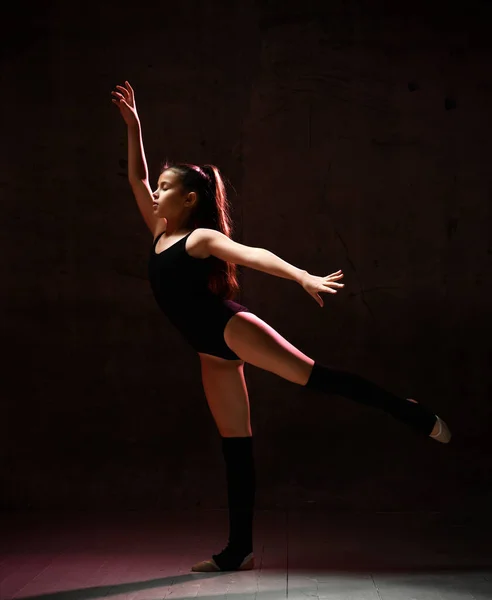 Νεαρή γυμνάστρια σε μαύρο αθλητικό σώμα και με ειδικό πάνω μέρος στέκεται και κάνει γυμναστική ποζάρουν σε σκούρο φόντο — Φωτογραφία Αρχείου