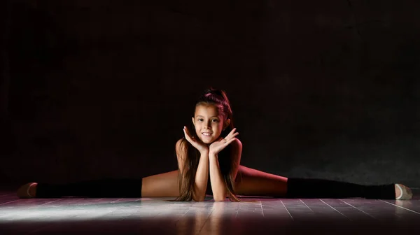 Young smiling girl gymnast in black sport body and special footwear sitting in twine over dark background — ストック写真