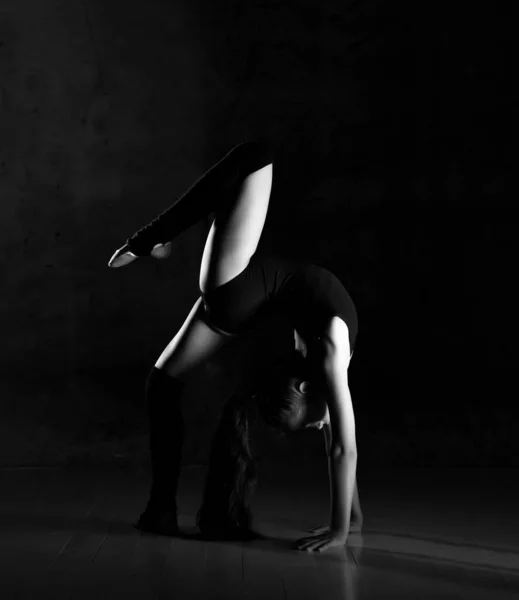 Young girl gymnast in black sport body and special footwear standing in bridge pose and keeping balance over dark background — Stock Photo, Image