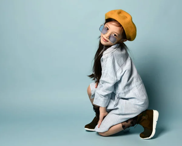 Niña asiática en gafas de sol, vestido de camisa, boina marrón, botas. Sonriendo, posando sentado de lado sobre fondo azul. De cerca. — Foto de Stock