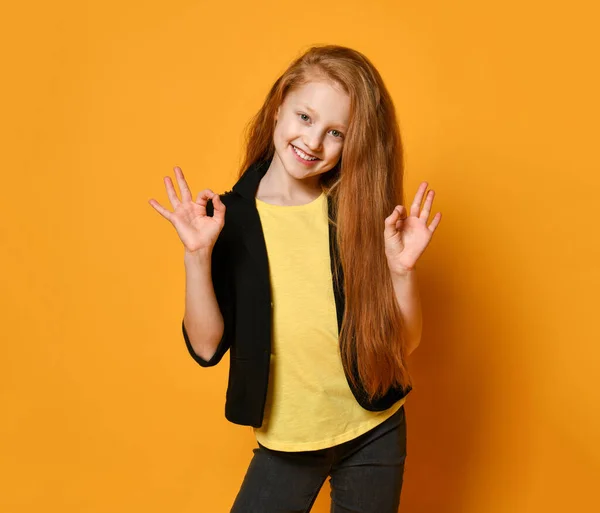 Ginger tienermeisje in zwarte jas, broek, geel t-shirt. Ze glimlacht, toont een goed teken, poseert op oranje achtergrond. Sluiten. — Stockfoto