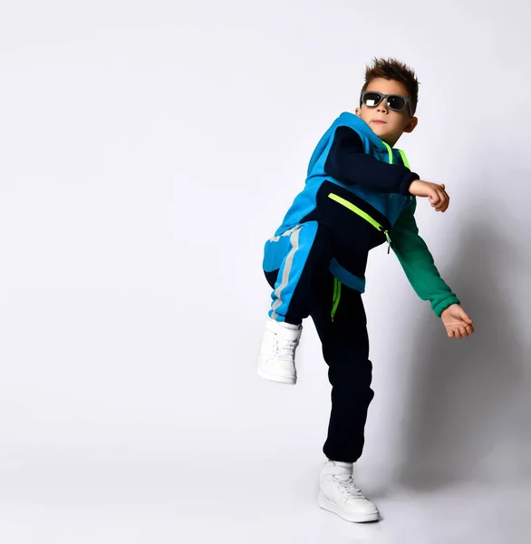 Mały dzieciak w okularach przeciwsłonecznych, kolorowym stroju sportowym i tenisówkach. Wykonuje kopnięcie w nogę, pozując samotnie na białym — Zdjęcie stockowe